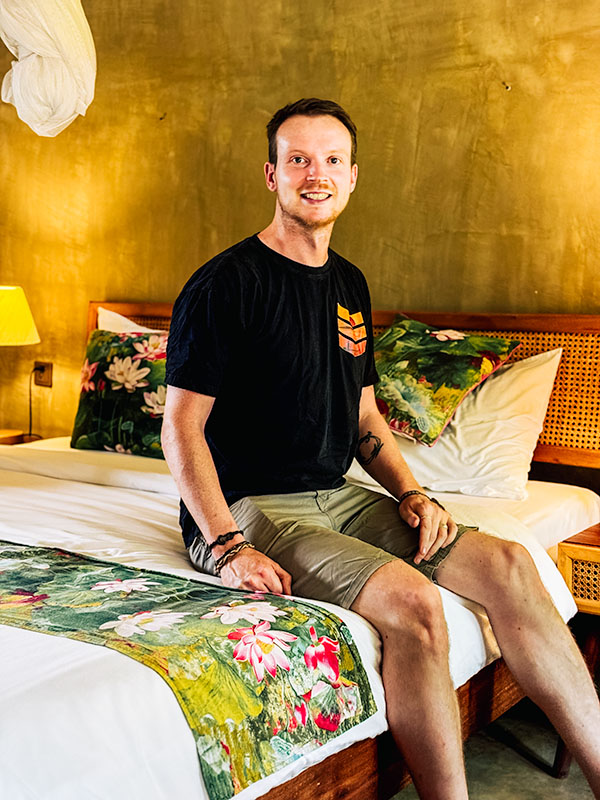 Matt sitting on a bed in a hotel in Cambodia with a floral bedspread