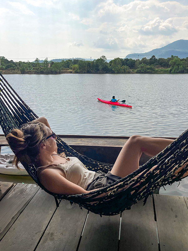 Katie lying in a hammock overlooking Kampot River as a red kayak paddles by