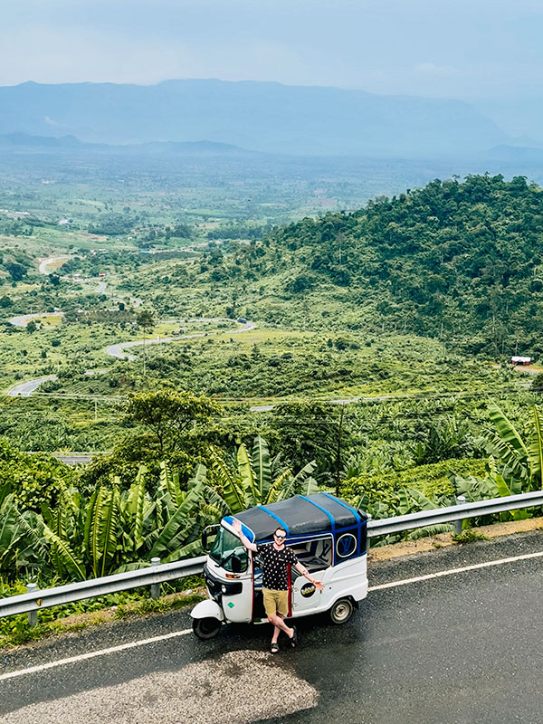 Matt standing in front of a white tuktuk (with TukTuk Rental branding) with winding road and jungle in the background